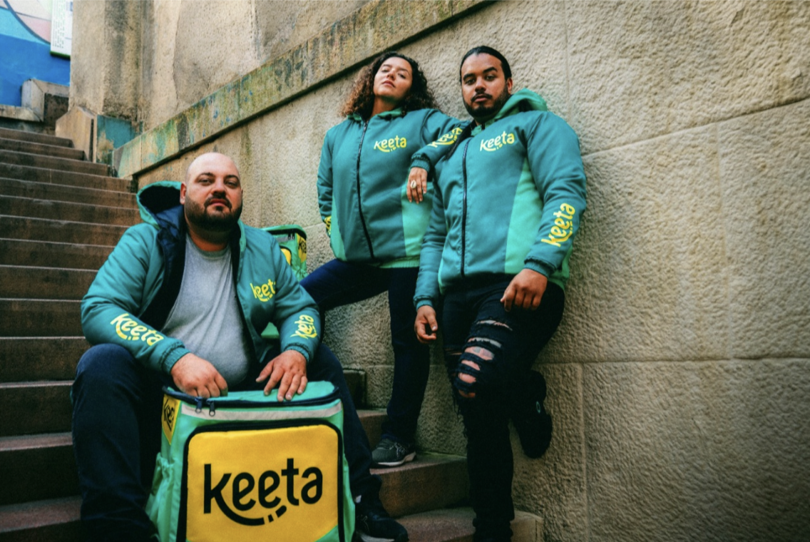 Três entregadores parceiros da Keeta usando jaquetas da plataforma posam em uma escadaria ao lado de uma mochila de entregas com a marca Keeta.