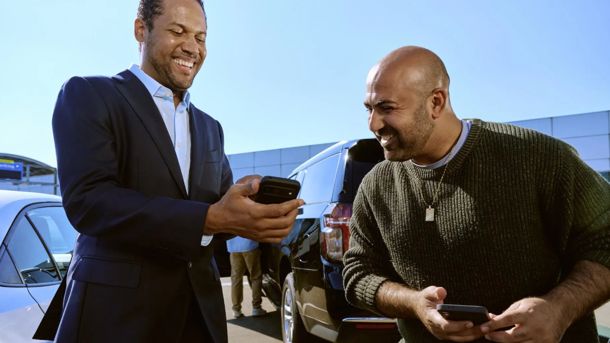 Dois homens sorriem enquanto observam um celular em uma área de estacionamento, com veículos ao fundo.