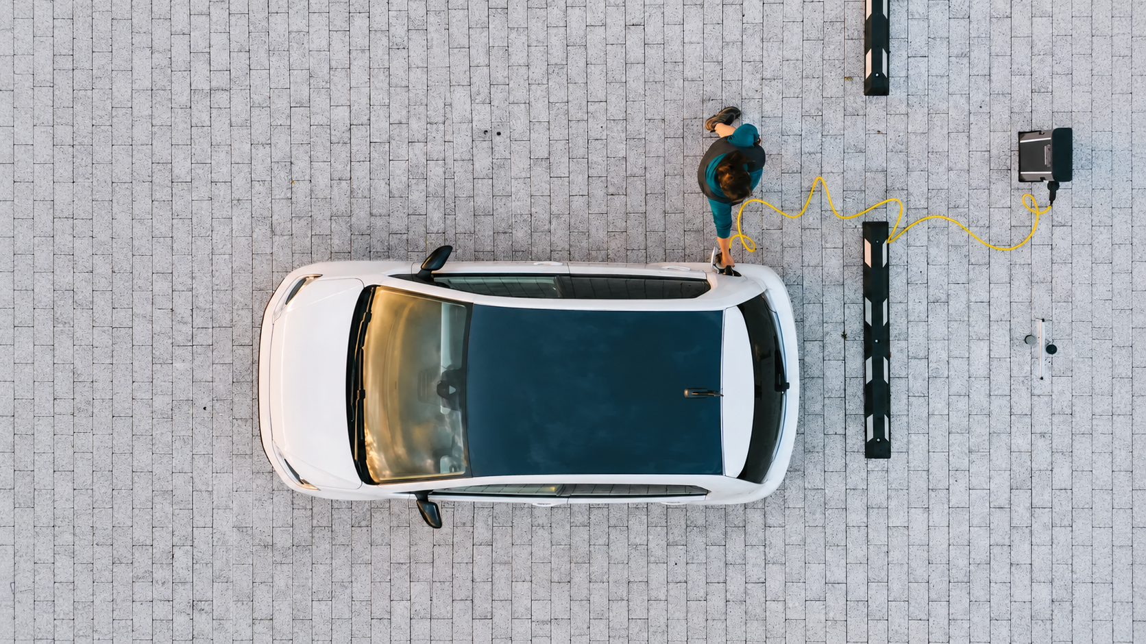 Vista aérea de um carro elétrico branco conectado a uma estação de recarga, com uma pessoa ao lado do veículo.