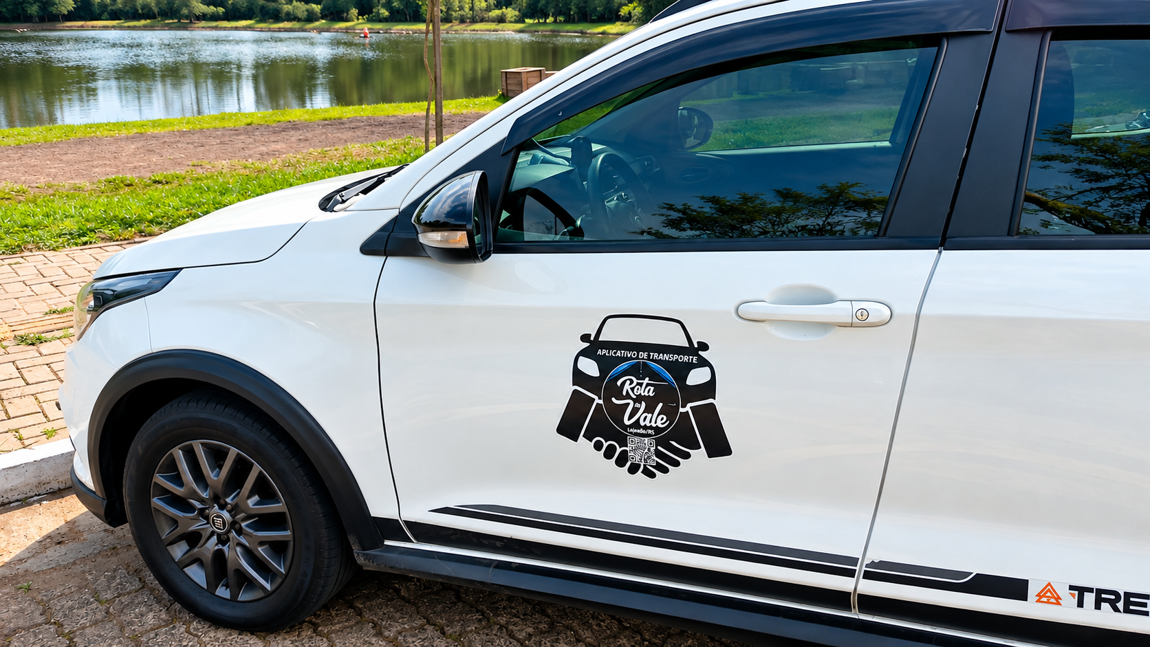 Carro branco com identificação do aplicativo de transporte Rota do Vale estacionado próximo a um lago.