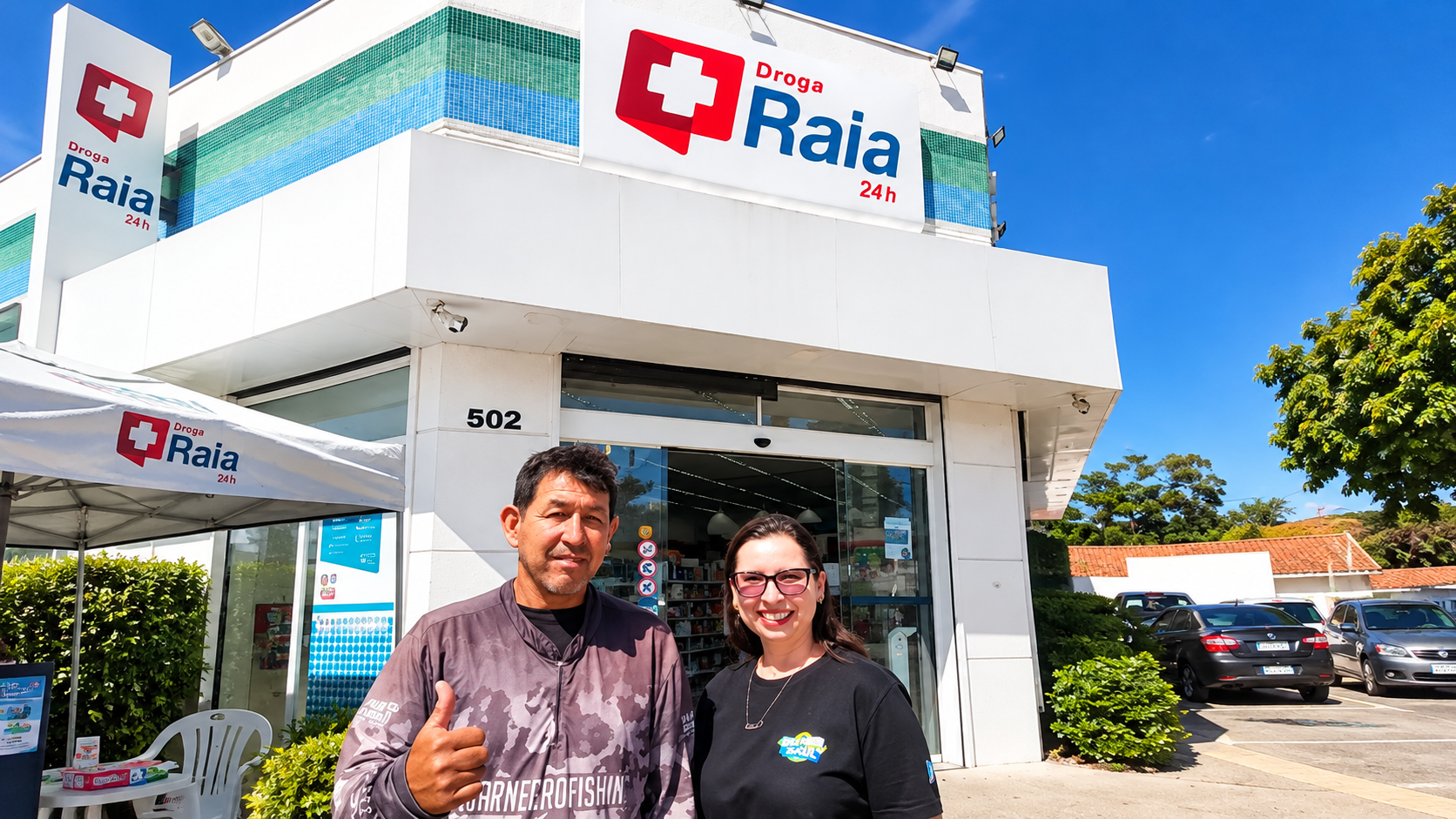 =Homem fazendo sinal de positivo ao lado de mulher com óculos, em frente a uma unidade da Droga Raia 24 horas, com fachada branca e logotipo da farmácia.