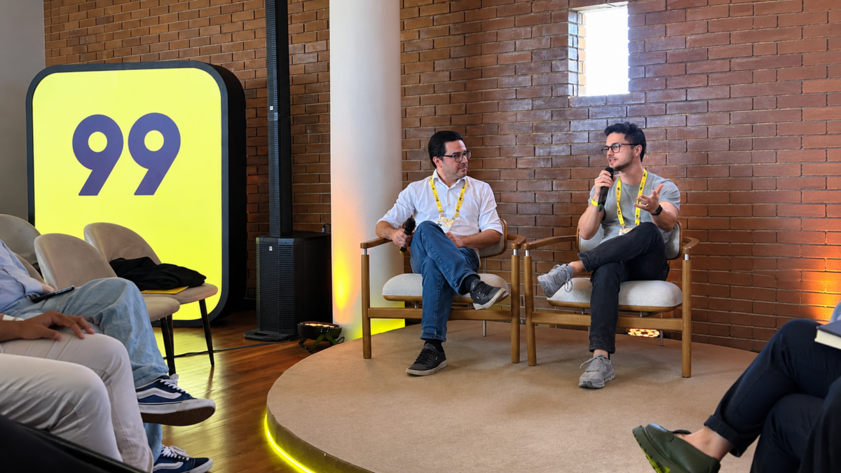Dois executivos participam de um painel da 99, sentados em poltronas, enquanto um deles fala ao microfone durante o evento.