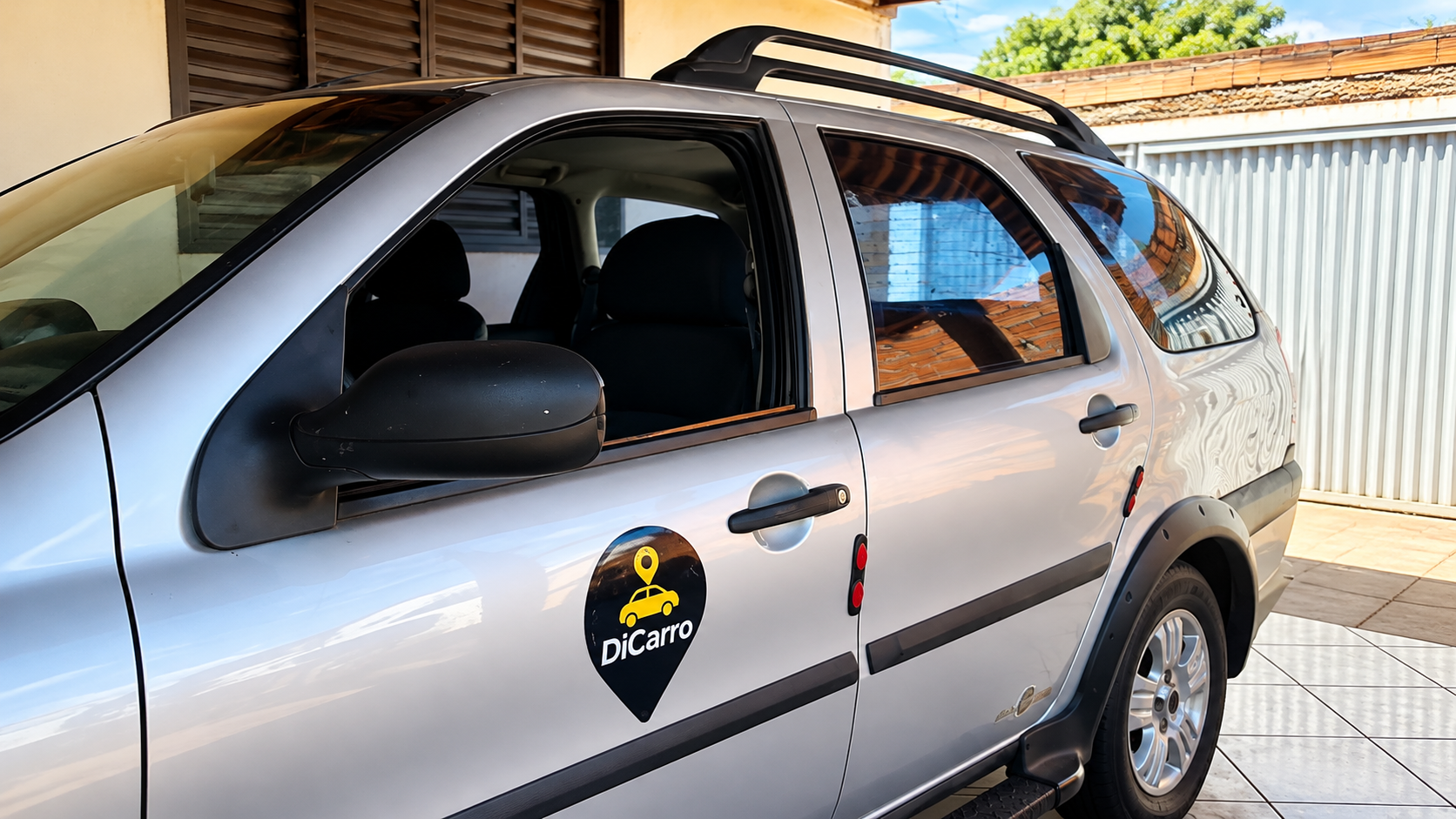 Veículo prata com adesivo da DiCarro Mobilidade Urbana na porta lateral, estacionado em garagem residencial coberta, com piso de cerâmica e portão branco ao fundo.