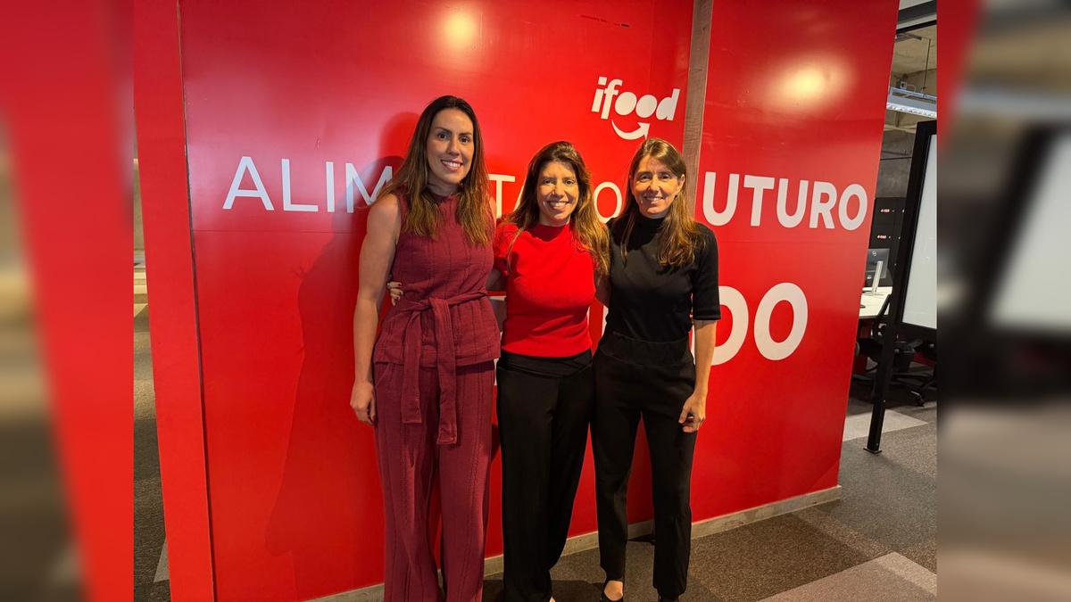 Três mulheres sorridentes posam lado a lado em frente a um painel vermelho com o logotipo do iFood e a frase parcialmente visível “Alimentando o Futuro”.