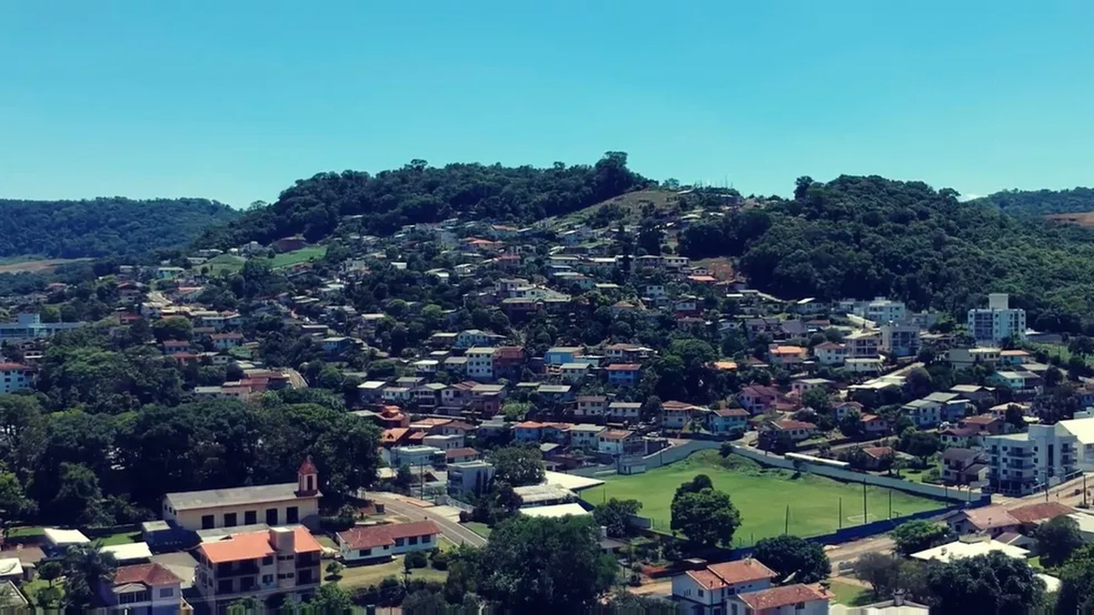 Vista panorâmica de bairro residencial com casas distribuídas entre morros arborizados, campo verde e igreja em primeiro plano.
