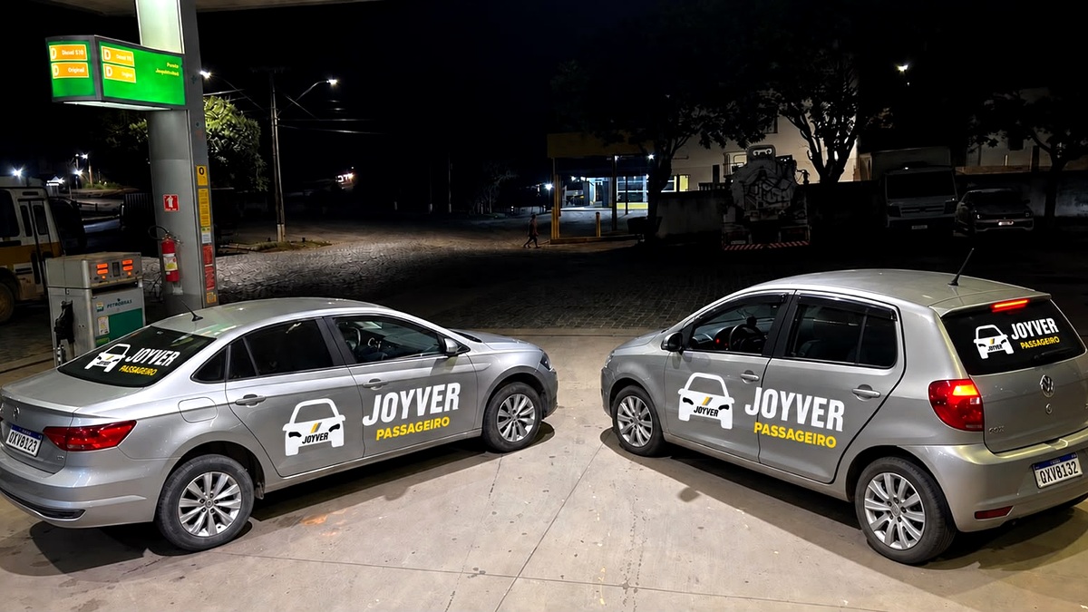Dois carros prata identificados com a marca Joyver Passageiro, estacionados lado a lado em um posto de combustíveis durante a noite.