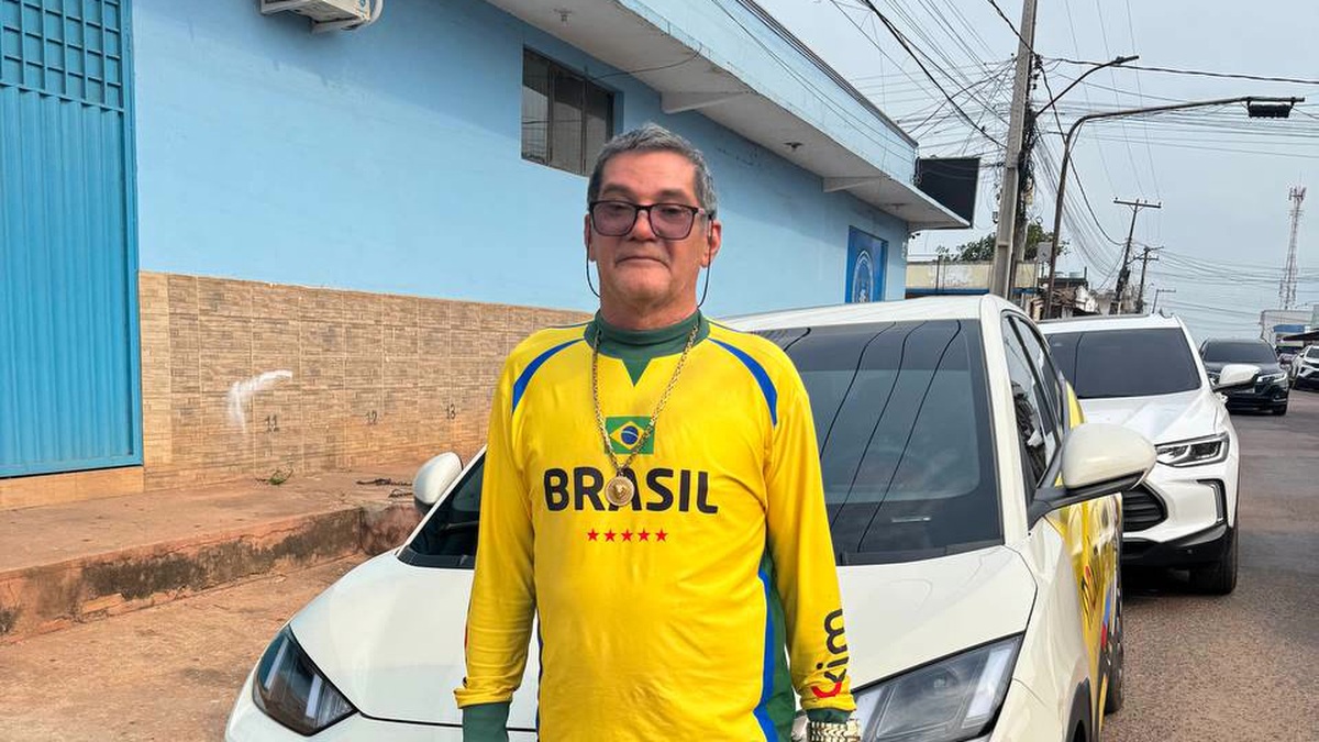 Motorista de aplicativo veste camiseta amarela do Brasil e posa em frente a um carro branco estacionado em uma rua.