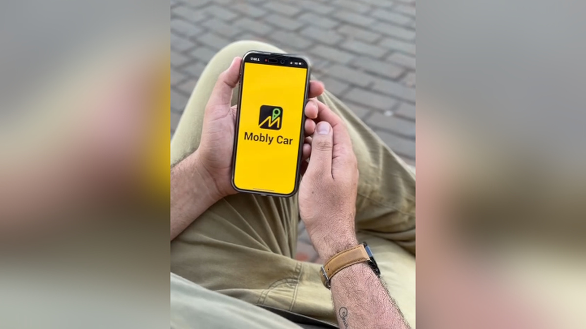 Pessoa sentada segura smartphone com a logomarca Mobly Car em tela amarela.